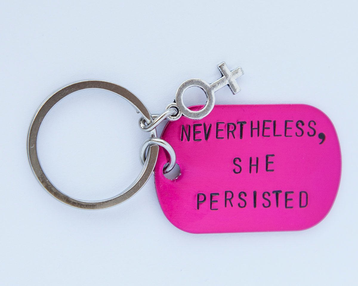 A pink feminist keychain with the phrase 'Nevertheless She Persisted' hand-stamped on a lightweight aluminum tag, attached to a durable stainless steel ring.